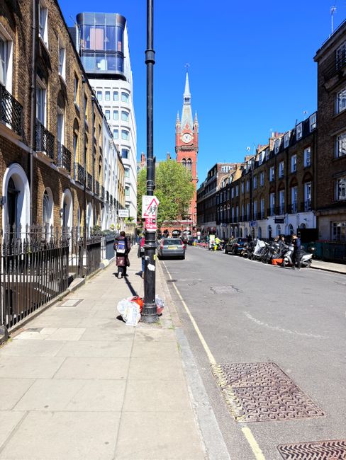 A straight, urban street scene during daytime showing a narrow pavement lined with a mix of traditional brick terraced houses and modern multi-story buildings, including a distinctive glass-fronted structure at the far end. The pavement features pedestrians, some carrying boxes or bags, and a black metal railing separating sidewalk from residential properties. A tall lamppost stands near the centre of the image, with a partially visible sign attached to it, and a plastic bag resting at its base. Parked cars and scooters are visible along the curb on the right side of the street. In the background, a prominent red-brick clock tower with a pointed spire rises above the surrounding buildings, with lush green trees interspersed among the structures. The scene is well-lit with bright sunlight and a clear blue sky, illustrating a typical day for house removals or furniture transport in Kennington, where logistical considerations such as narrow roads and access issues are a common aspect of home relocation projects, as handled by Removal Companies Kennington.