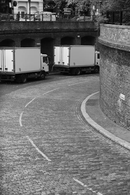 A black and white photograph depicts two medium-sized moving trucks parked on a cobbled street adjacent to a brick building, which curves along the roadside. The trucks are positioned side by side, facing towards a brick archway under an elevated area with a metal railing and trees visible on the upper level. The street has dashed white lines indicating parking or loading zones, and a narrow sidewalk runs along the base of the brick wall on the right side of the image. The environment appears to be part of an urban residential or commercial area, suitable for home relocation or furniture transport activities carried out by Removal Companies Kennington, as they assist with packing and loading processes in such settings.
