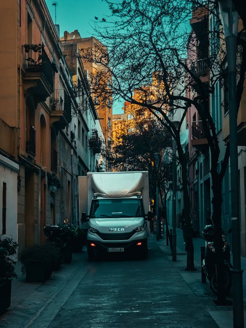 A moving van with a white cargo area and a black cab is backed into a narrow city street designed for pedestrian access, with tall, multi-storey residential buildings on either side. The van is positioned in front of an open doorway, with the rear doors slightly ajar, indicating the loading or unloading process. Several cardboard boxes and wrapped furniture items are visible on the pavement around the van, suggesting a home relocation. The street is lined with black metal bollards, some of which support bicycles, and there are leafless trees overhead, casting shadows on the pavement. The surrounding environment is illuminated by natural daylight, with warm hues from the buildings and cool tones from the street scene. The image exemplifies the logistical aspect of removals services, highlighting furniture and packing materials being moved during a house relocation, as provided by Removal Companies Kennington.
