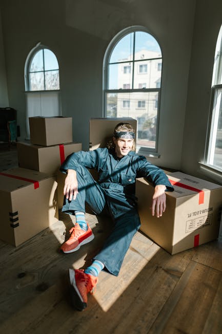 A young man wearing a blue jumpsuit, a patterned headband, and bright orange sneakers is sitting on a wooden floor inside a room with large arched windows that let in natural daylight. He is surrounde