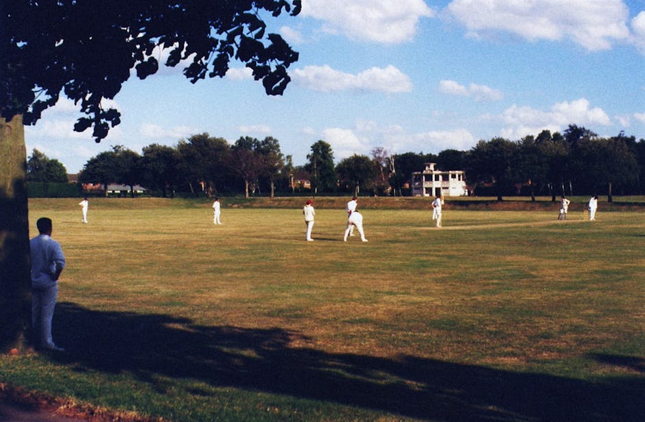 A wide view of a cricket practice field on a sunny day, with several players dressed in white cricket uniforms spaced evenly across the grassy area, preparing for or participating in training. In the foreground, a person wearing a blue blazer and white trousers stands under the shade of a tree, observing the activity. The field is surrounded by trees and residential buildings in the background, with a partly cloudy sky overhead. This scene may be associated with outdoor sports activities or team practice sessions, with the open grassy space and equipment such as cricket bats and balls possibly present. The image, related to house removals or relocation services, is captured at a distance but highlights the outdoor environment and movement context in the vicinity of a sports ground, which could be on or near a residential property managed by Removal Companies Kennington.