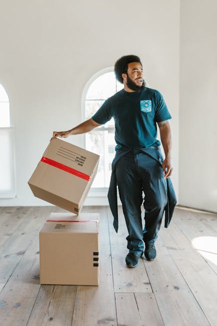 A man with dark curly hair and a beard is inside a well-lit room with a large arched window in the background. He is wearing a navy blue t-shirt and dark trousers, and has a sweater tied around his waist. He is lifting a medium-sized cardboard box with red tape around it, preparing to place it onto a similar box on the wooden floor. The room has plain white walls and light wooden flooring, suggesting a home environment during a house removal process. This image depicts the packing and furniture transport aspects of a home relocation, with the man engaging in the loading or unloading stage of moving services, as part of the operations undertaken by Removal Companies Kennington. The setting is clear and detailed, illustrating typical packing materials and the moving process involved in house removals and furniture transport.