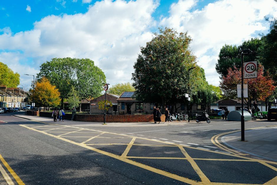 A street scene in a residential area featuring a small brick building surrounded by tall trees with green, yellow, and orange leaves, indicating autumn. The intersection has yellow grid markings on the asphalt for traffic control, and there are pedestrians walking along the pavements. Several bicycles are parked against a metal railing near the building, and a few motorcycles and cars are visible on the street. The sky is partly cloudy with patches of blue, and there are two street signs, one indicating a 20 mph zone, positioned near black lampposts. This setting depicts a typical neighbourhood environment, which could be involved in home relocation or furniture transport services. Removal Companies Kennington may utilise such areas for loading or unloading during house removals and moving logistics.
