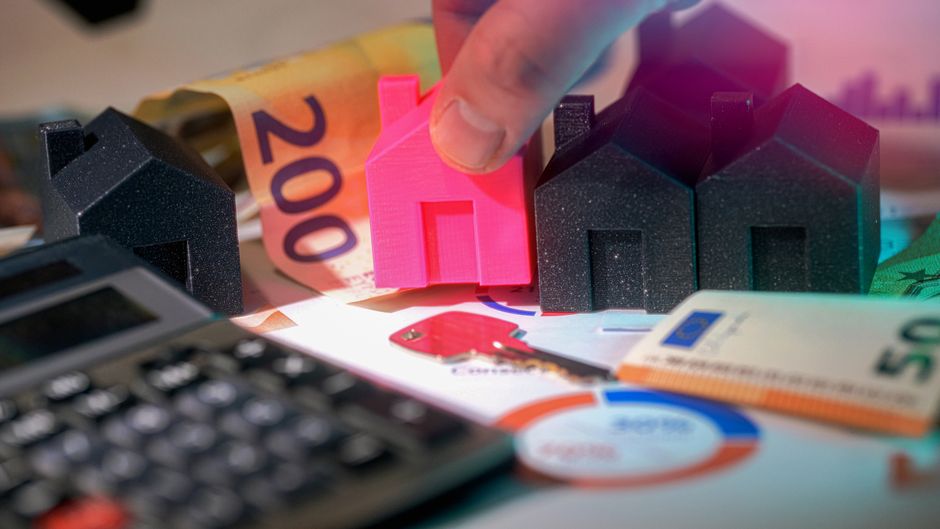A close-up image showing a person's hand placing a small, pink, 3D-printed house model among other miniature black house models on a cluttered surface. Surrounding the models are various financial doc