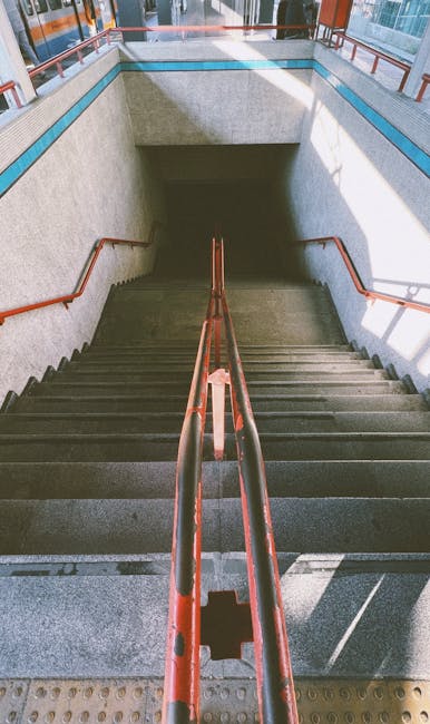 The image shows a concrete staircase inside a building, viewed from the top looking down, with red metal handrails on both sides and a textured yellow tactile paving at the top edge. The stairs descend into a well-lit corridor, with partially visible surrounding walls and glass windows allowing natural light to illuminate the area. This setting represents the interior of a transport hub or public building, where staircases are used for access between floors. Such stair access can present challenges during home relocation or furniture transport, particularly when maneuvering large or bulky items. In the context of house removals, professionals from Removal Companies Kennington may need to navigate narrow staircases like this, using appropriate moving equipment such as trolleys, straps, and blankets to ensure safe handling of furniture and boxes during packing and moving processes within buildings with limited stair access.