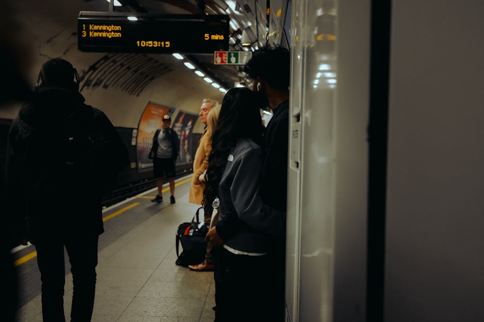 Inside Kennington Station, several passengers are waiting on the platform near the edge, with some standing close to a white pillar and others further back. A digital display board overhead shows train information, including upcoming arrivals to Kennington and surrounding stations, with times and platform details. The platform surface appears clean with visible yellow safety lines along the edge. The lighting is artificial, typical of underground stations, creating a slightly dim atmosphere. In the background, there are advertisements on the curved station wall, enhancing the typical underground environment. This scene captures the typical moment during home relocation or furniture transport, where passengers wait before boarding, potentially supporting the logistics and booking processes of removals and moving services, as carried out by [COMPANY_NAME].