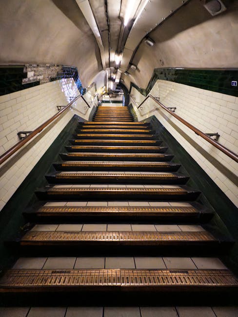 The image shows a concrete staircase inside a building, viewed from the top looking down, with red metal handrails on both sides and a textured yellow tactile paving at the top edge. The stairs descend into a well-lit corridor, with partially visible surrounding walls and glass windows allowing natural light to illuminate the area. This setting represents the interior of a transport hub or public building, where staircases are used for access between floors. Such stair access can present challenges during home relocation or furniture transport, particularly when maneuvering large or bulky items. In the context of house removals, professionals from Removal Companies Kennington may need to navigate narrow staircases like this, using appropriate moving equipment such as trolleys, straps, and blankets to ensure safe handling of furniture and boxes during packing and moving processes within buildings with limited stair access.