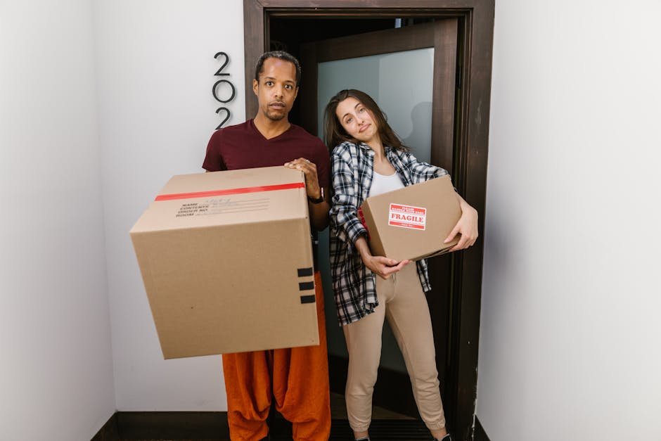 A man and a woman stand inside a residential doorway, each holding cardboard boxes for house removal. The man, dressed in a maroon t-shirt and orange trousers, holds a large box with red and black tape, while the woman, wearing a plaid shirt and beige trousers, cradles a smaller box labeled 'FRAGILE'. They appear to be preparing for a home relocation, with their expressions neutral and focused. The background includes the doorframe, and the surroundings suggest an indoor hallway or entryway. The scene illustrates the loading process involved in professional furniture transport and packing for removals, as provided by Removal Companies Kennington, supporting house moving services in Kennington and the wider Lambeth area.