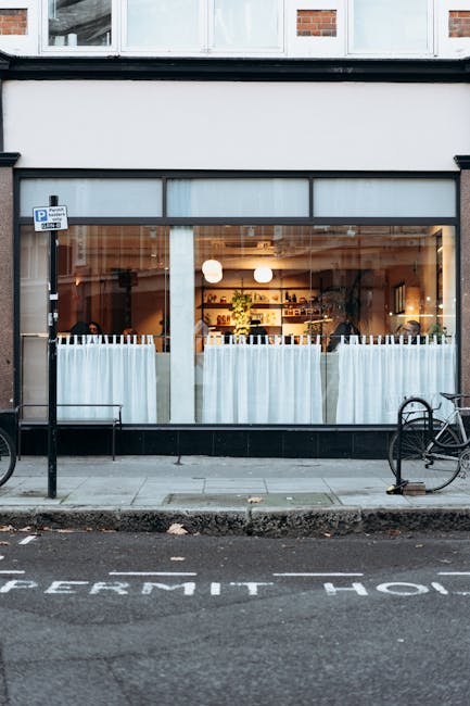 A storefront with large glass windows revealing the interior of a café or restaurant, illuminated by warm lighting and decorated with potted plants and shelving units. Outside, on the sidewalk, a black bicycle is parked against a metal railing, and a parking sign is visible above. The pavement features road markings indicating parking restrictions, and the building's exterior includes a brick facade above the shopfront. This scene relates to house removals or furniture transport processes, which may involve moving items into or out of commercial premises, supported by companies like Removal Companies Kennington, operating within professional relocation and logistics services.