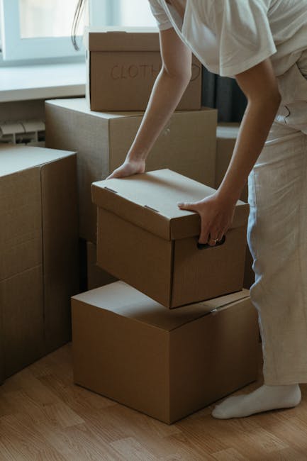 A man and a woman stand inside a residential doorway, each holding cardboard boxes for house removal. The man, dressed in a maroon t-shirt and orange trousers, holds a large box with red and black tape, while the woman, wearing a plaid shirt and beige trousers, cradles a smaller box labeled 'FRAGILE'. They appear to be preparing for a home relocation, with their expressions neutral and focused. The background includes the doorframe, and the surroundings suggest an indoor hallway or entryway. The scene illustrates the loading process involved in professional furniture transport and packing for removals, as provided by Removal Companies Kennington, supporting house moving services in Kennington and the wider Lambeth area.