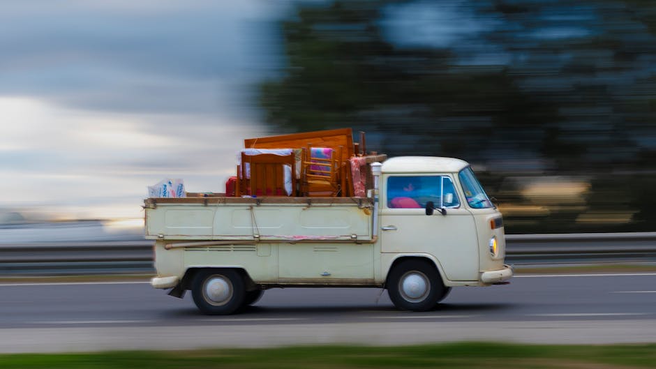 A vintage cream-coloured van with a compact, rounded shape, typical of older European utility vehicles, is shown in motion on a street, captured with a blurred background indicating speed. The van's cargo area is loaded with various household items, including wooden chairs, a table, and plastic and cardboard boxes, some wrapped in protective materials like fabric or plastic sheets. The furniture appears to be securely stacked and arranged to prevent movement during transit. The vehicle is part of a home relocation or furniture transport process associated with professional removals, possibly managed by company services such as Removal Companies Kennington. The scene depicts the loading or transportation phase in a moving service, highlighting the importance of careful packing and secure vehicle loading on a moving day, with an emphasis on efficiency and safety during the loading process.
