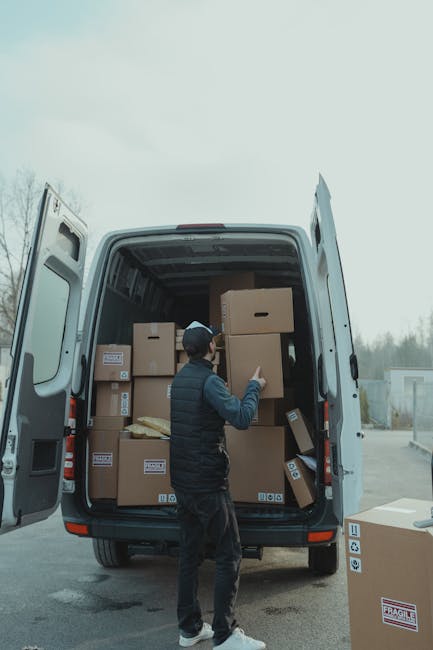 A man dressed in a black jacket, dark trousers, and a cap is standing in front of an open white delivery van with its rear doors fully open. Inside the van, there are several cardboard boxes of various sizes, some with labels and barcodes, stacked on top of each other and secured in the cargo area, which is illuminated by natural light from outside. The man is in the process of arranging or moving the boxes, possibly part of a home relocation or furniture transport operation. The van is parked on a paved surface, and a few more boxes, including another labeled 'fragile,' are placed on the ground nearby. The background shows a cloudy sky and a distant view of trees and industrial buildings, consistent with an urban or suburban moving environment. This scene exemplifies the loading process during a professional removal or moving service, illustrating standard packing and furniture transport procedures facilitated by Removal Companies Kennington.