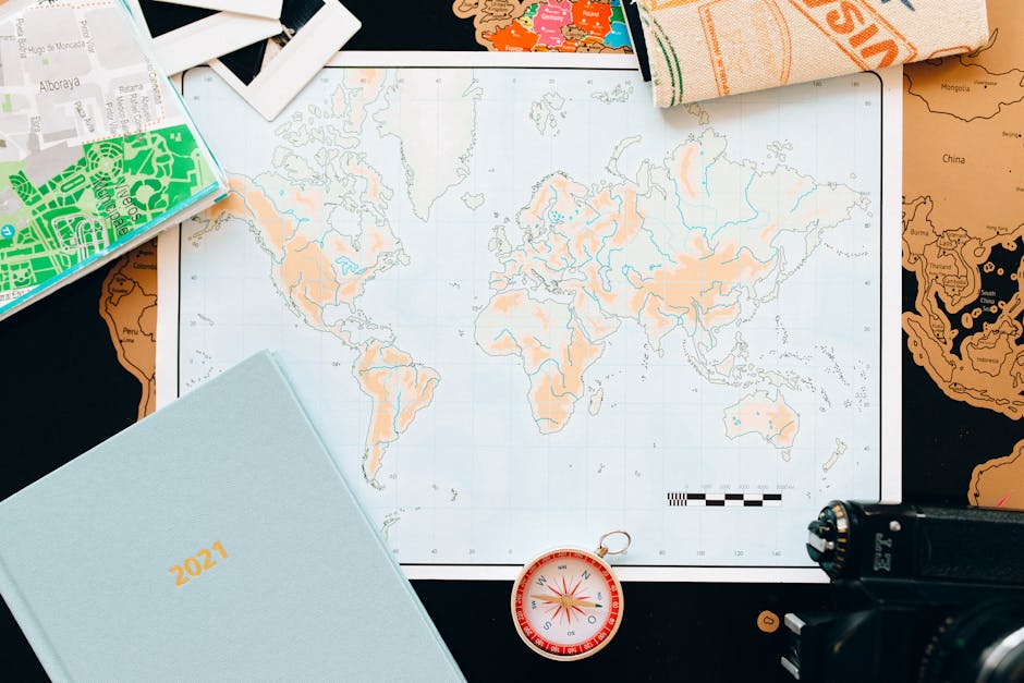 A flat-lay arrangement of travel and planning items on a black surface, including an open world map with detailed continent outlines and country borders, which is partially covered by various objects. There is a closed light blue 2021 planner positioned in the bottom left corner. Near the center, a small, round compass with a red and white face and a metal ring is visible, suggesting navigation or travel planning. To the top left, a partially visible cardboard box with green and white abstract print is present, and to the top right, a folded towel or cloth with orange, white, and green patterns is seen. Additionally, a black camera is placed in the lower right corner, indicating documentation or photography related to home relocation or logistics. The overall scene suggests planning or mapping for an international move or travel, consistent with services offered by Removal Companies Kennington for house removals and relocation logistics.