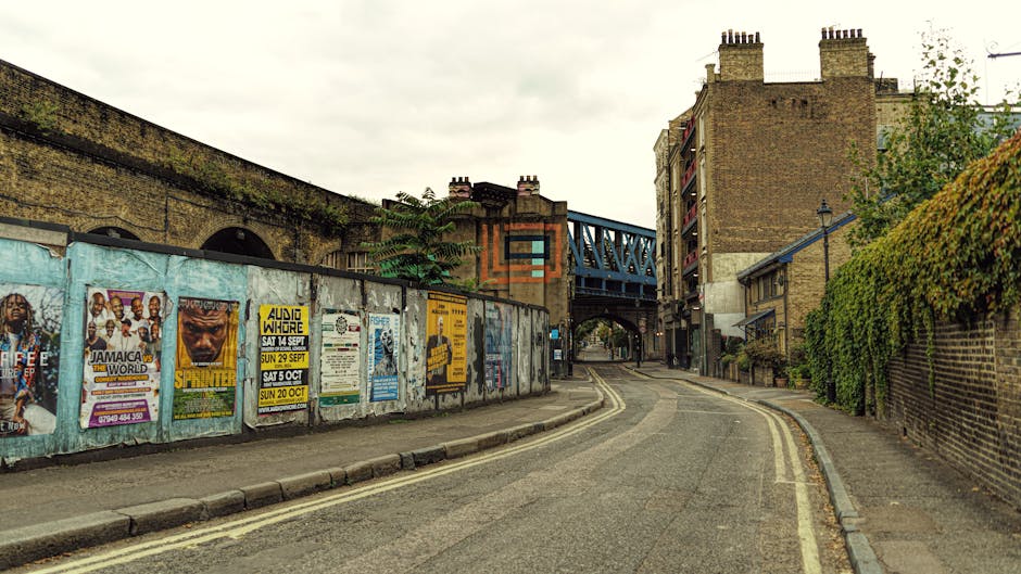 A narrow urban street scene showing a curved asphalt road with yellow double lines on the edge, leading towards an underpass and a blue railway bridge in the background. On the left side of the street, there is a graffiti-covered metal fence displaying various colorful posters advertising local events and performances, including music and comedy shows. The fence is slightly worn with some rust and peeling paint. Behind the fence, an old brick wall and a large building with ventilation pipes and a chimney are visible, indicating an industrial or commercial area. On the right side, there is a brick wall partially covered with green ivy climbing up the surface, along with residential buildings facing the street, some with small balconies and windows. The scene is overcast with diffused natural light, typical of a typical city environment during daytime, connected to house removals and logistics, illustrating an urban setting where furniture or boxes might be loaded into a van for home relocation by Removal Companies Kennington.