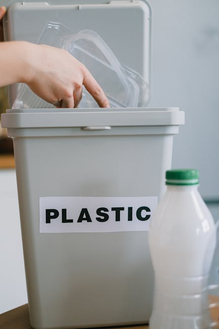 A hand placing a clear plastic bottle into a light grey plastic recycling bin labeled 'PLASTIC' with a white background. In the foreground, a partially visible white plastic bottle with a green cap is situated on a wooden surface. The setting appears to be indoors, possibly within a home or moving environment, as part of the packing and waste disposal process associated with house removals. The scene reflects an organized approach to recycling during home relocation, coordinated by Removal Companies Kennington, with the bin positioned near an open space or doorway, indicating ongoing preparation for a move or clearance. The lighting is neutral and natural, highlighting the clarity of the plastic items and the cleanliness of the area, underscoring proper waste separation for eco-friendly disposal during furniture transport or packing routines.
