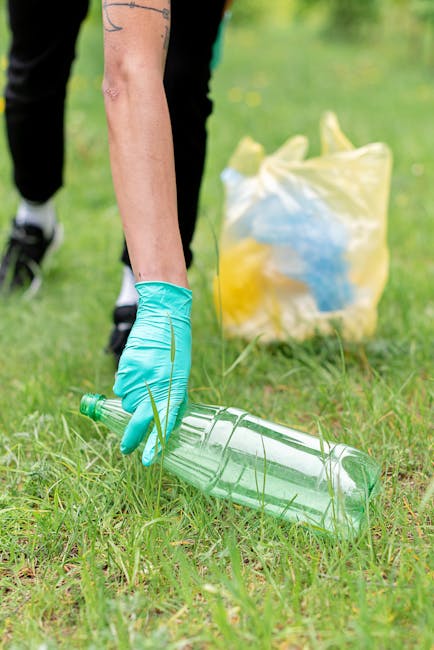 A person wearing a blue disposable glove is bending down and picking up a clear plastic bottle from a grassy area outside a property during a home relocation or packing process. In the background, there is a partially visible yellow and white plastic bag containing waste or donation items, and a blue object inside the bag. The scene appears to take place outdoors, possibly near a doorway or driveway used for loading belongings onto a removal vehicle. The image emphasizes waste disposal and recycling as part of the moving process, aligning with the services of Removal Companies Kennington, who assist with packing, waste management, and furniture transport during house removals.