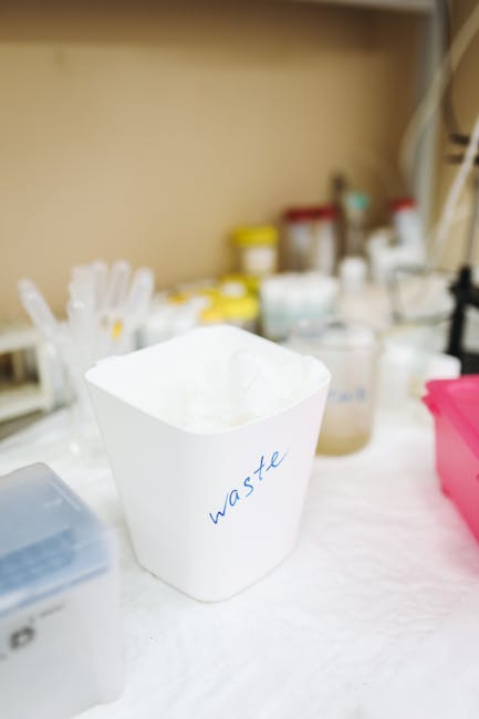 A hand placing a clear plastic bottle into a light grey plastic recycling bin labeled 'PLASTIC' with a white background. In the foreground, a partially visible white plastic bottle with a green cap is situated on a wooden surface. The setting appears to be indoors, possibly within a home or moving environment, as part of the packing and waste disposal process associated with house removals. The scene reflects an organized approach to recycling during home relocation, coordinated by Removal Companies Kennington, with the bin positioned near an open space or doorway, indicating ongoing preparation for a move or clearance. The lighting is neutral and natural, highlighting the clarity of the plastic items and the cleanliness of the area, underscoring proper waste separation for eco-friendly disposal during furniture transport or packing routines.